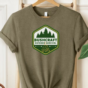 以下が含まれることがあります： キャンプファイヤー、木々、そして「Bushcraft Outdoor Survival」というテキストが書かれた六角形のワッペンが付いた緑色のTシャツです。