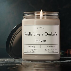 May include: A glass jar candle with a beige label that reads "Smells Like a Quilter's Haven". The label also includes details like "9 oz / 255 g", "Soy Wax", "Made in USA", "Cotton Wick", "50 Hour Burn Time", and "Since 2025".