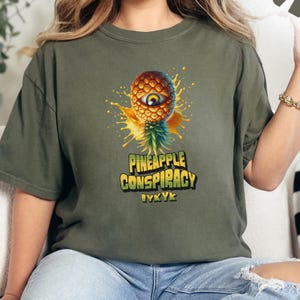 以下が含まれることがあります： パイナップルのグラフィックと「Pineapple Conspiracy」と「IVYK」のテキストが黄色と緑の文字で書かれた緑色のTシャツです。