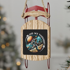 May include: A wooden Christmas ornament shaped like a sled with a black sign that says "Did We Though?" and a cartoon astronaut in space.