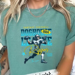 Può includere: Una maglietta verde acqua con un design grafico di un giocatore di football americano. Il design include il testo "Fantasy Football", "Doghouse League", "2024", "Rippers" e "League Champs".