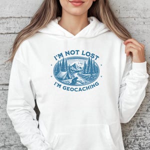 Puede incluir: Sudadera con capucha blanca con un estampado gráfico azul que dice "No estoy perdido, estoy haciendo geocaching". El gráfico presenta una escena de montaña con una tienda de campaña, árboles y una persona con una mochila.