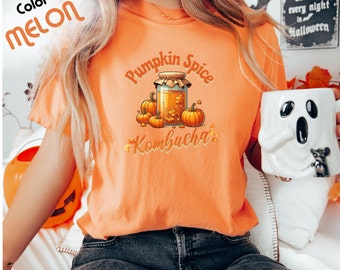 May include: A woman wearing a bright orange t-shirt with the text "Pumpkin Spice Kombucha" and a graphic of a jar of kombucha with pumpkins. The shirt is a light orange color.