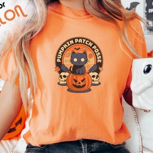 May include: Orange t-shirt featuring a black cat perched on a jack-o'-lantern. The design reads "Pumpkin Patch Posse" in a circle around the cat and pumpkin.