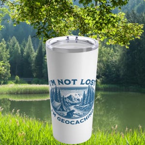 May include: White stainless steel tumbler with a blue and white graphic of a forest scene with a tent, a river, and a bear. The text "I'm not lost I'm geocaching" is printed on the tumbler.