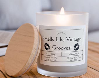 Puede incluir: Un tarro de vela de vidrio esmerilado con una tapa de madera. La etiqueta dice "Smells Like Vintage Grooves!" e incluye detalles como "9 oz / 255 g", "Cera de soja de coco", "Hecho en EE. UU.", "Mecha de madera", "Tiempo de combustión de 50 horas" y "Desde 2025".