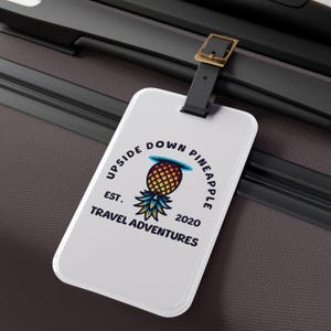以下が含まれることがあります： 黒と黄色のパイナップルのデザインが施された白い荷物タグ。テキストには「Upside Down Pineapple Est. 2020 Travel Adventures」と書いてあります。