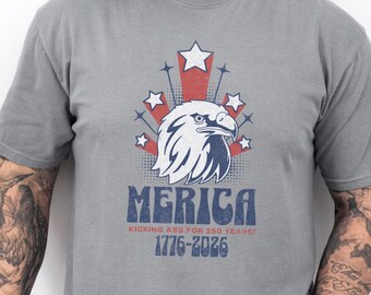 May include: Gray t-shirt featuring a patriotic design with an eagle, stars, and the word "MERICA." The shirt includes the text "Kicking Ass for 250 Years!" and the dates 1776-2026.