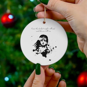 Puede incluir: Adorno navideño de cerámica blanca con una ilustración en blanco y negro de una mujer con los ojos cerrados. El adorno tiene el texto "Incluso la noche más oscura terminará y el sol saldrá."
