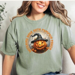 May include: A green t-shirt with a graphic of a pumpkin wearing a witch's hat and the text "Witching Hour With Pumpkin Power".