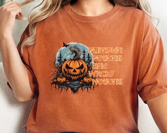 May include: Orange t-shirt with a graphic of a jack-o'-lantern in front of a moon and spooky trees. The text "Pumpkin Potions and Witchy Notions" is printed on the shirt.