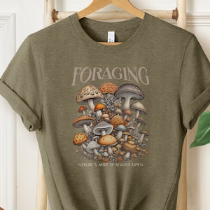 May include: A green t-shirt with a graphic of a cluster of mushrooms in various colors and shapes. The text "FORAGING" is above the mushrooms and "NATURE'S AISLE IS ALWAYS OPEN" is below the mushrooms.