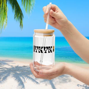 以下が含まれることがあります： 竹製の蓋とパイナップルのグラフィックが描かれた黒と白の「IYKYK」ロゴが付いた透明なガラス製の再利用可能な缶。ガラス製のストローが缶に挿入されています。
