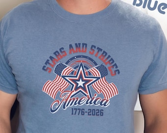 May include: A blue short-sleeved t-shirt with a patriotic design. The graphic includes the text "STARS AND STRIPES America 1776-2026" with a star and American flags. The shirt is a solid blue color.