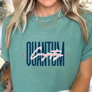 Puede incluir: Una camiseta azul turquesa con el texto "QUANTUM Computing QUANTUM" en azul marino y rojo.