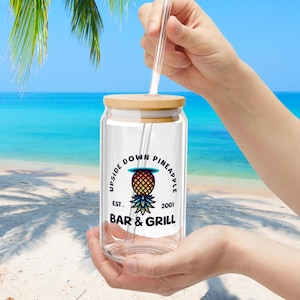以下が含まれることがあります： 木製の蓋とストローが付いた透明なガラス瓶。瓶には「Upside Down Pineapple Bar & Grill Est. 2001」という文字とパイナップルのグラフィックが貼られたステッカーがあります。
