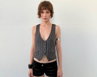 May include: A charcoal gray pinstriped vest featuring a deep V-neckline, three front buttons, and two small decorative pockets at the waist, paired with black shorts. The vest has a tailored fit with a pointed hemline at the center front.