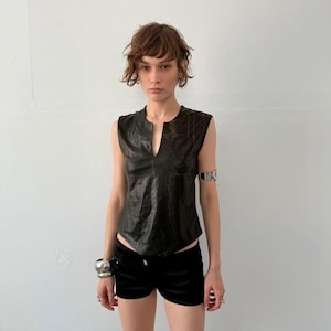 May include: A sleeveless black top with a subtle snakeskin texture and a deep V-neckline, paired with black shorts. The outfit is accessorized with a silver arm cuff on the left bicep and a thick silver bracelet on the right wrist.