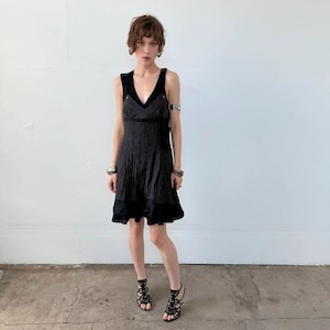 May include: A black sleeveless dress featuring a V-neckline, a textured bodice with a wrap-style front, and a solid black hem panel. The dress has a knee-length skirt and is paired with black strappy high-heeled sandals and silver-toned jewelry.