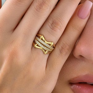 May include: A gold-colored ring with a star-like design, featuring two bands of small, clear stones. The ring is worn on a finger, with the hand partially covering the face. The ring's design is geometric and modern.