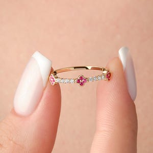 Könnte beinhalten: Ein zarter Goldring mit einer Reihe kleiner Diamanten und drei rosafarbenen Edelsteinen, die in Form eines Kleeblatts gefasst sind.