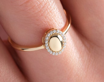 May include: A gold ring with a round, oval-shaped center stone surrounded by smaller diamonds. The ring is on a person's finger.
