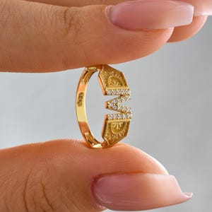 May include: A gold ring with the letter "M" encrusted with small, clear gemstones. The ring has a decorative sunburst design on the top and bottom. The ring is held between two fingers.