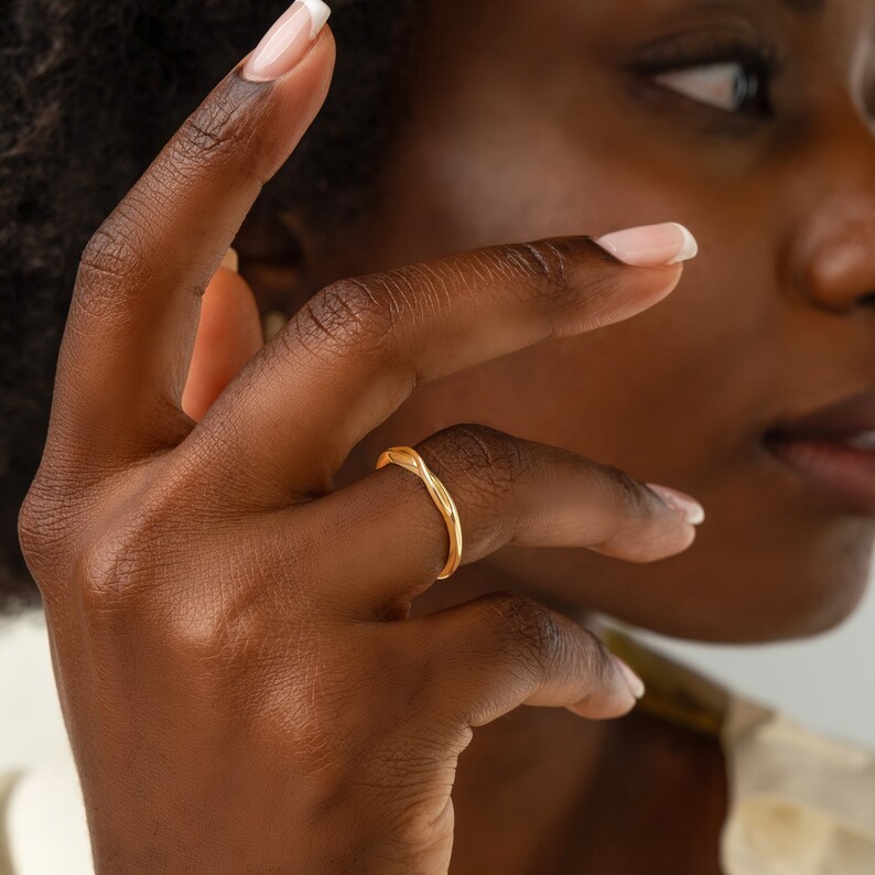 May include: A gold ring with a unique, wavy design. The ring is worn on the finger of a person with dark skin.