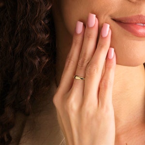 May include: A simple gold band ring with a rounded, slightly curved shape. The ring is worn on the ring finger of a person's left hand.