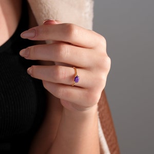 May include: A gold ring with a purple oval gemstone. The ring is on a person's finger.