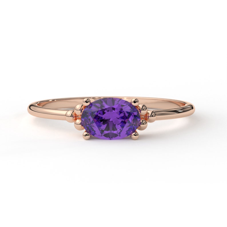 May include: A rose gold ring with a purple oval gemstone set in a delicate prong setting. The ring has a simple band and is accented with small beads on either side of the stone.