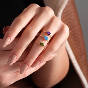 May include: Three gold rings with oval-shaped gemstones. The top ring has a purple gemstone, the middle ring has a blue gemstone, and the bottom ring has a green gemstone.