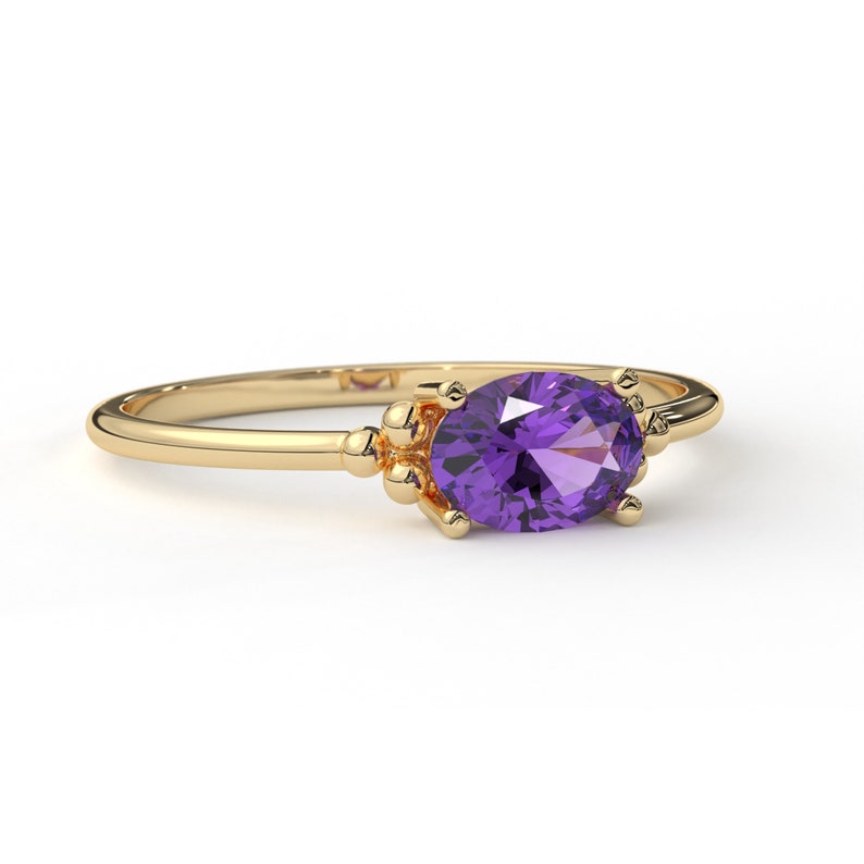 May include: A gold ring with a purple oval gemstone set in a delicate prong setting. The ring has a simple band with small gold beads on either side of the stone.