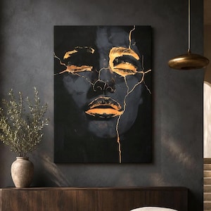 May include: A rectangular wall art piece featuring a dark, moody portrait of a face with closed eyes. The face is accented with bright gold, crack-like lines running across the forehead, eyes, and lips, creating a striking contrast against the black.