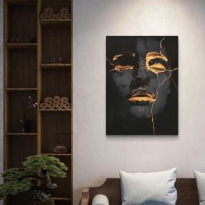 May include: A rectangular wall art print featuring a dark, monochromatic portrait of a face with closed eyes and lips. The image is accented with bright gold lines that resemble cracks running across the forehead, nose, and chin areas.