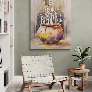 May include: A framed art print featuring a watercolor illustration of a broken pot with gold accents, and the text "You are not broken, you are becoming." The artwork hangs above a woven chair and wooden side table.