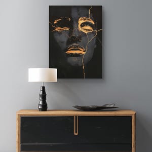 May include: A rectangular wall art print featuring a dark, shadowy human face with closed eyes, accented by metallic gold cracks running across the forehead, nose, and lips, displayed above a wooden sideboard with a black front and a table lamp.