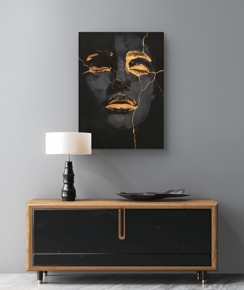 May include: A rectangular wall art print featuring a dark, shadowy human face with closed eyes, accented by metallic gold cracks running across the forehead, nose, and lips, displayed above a wooden sideboard with a black front and a table lamp.
