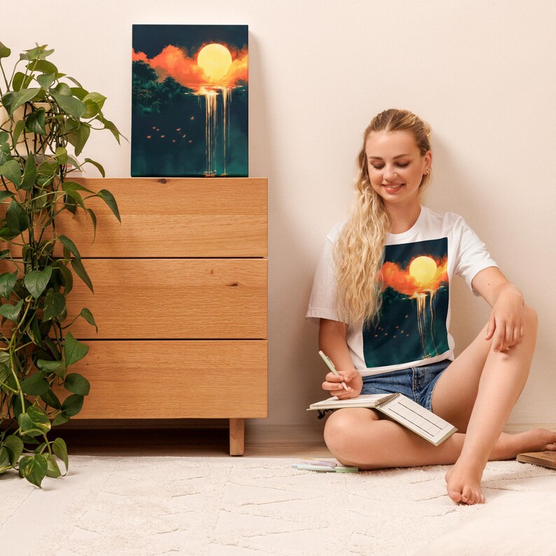 May include: A white t-shirt with a graphic print of a yellow sun setting over a landscape with a waterfall. The person wearing the shirt is sitting on the floor with a notebook and pen.