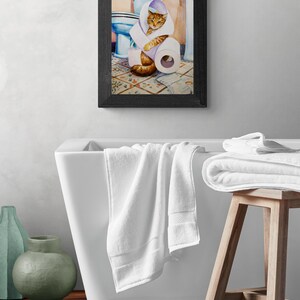 Peut inclure: Une peinture &agrave; l'aquarelle encadr&eacute;e repr&eacute;sente un chat roux envelopp&eacute; dans du papier toilette dans une salle de bain. La peinture est accroch&eacute;e au-dessus d'une baignoire blanche avec des serviettes blanches. Un tabouret en bois contient d'autres serviettes blanches.