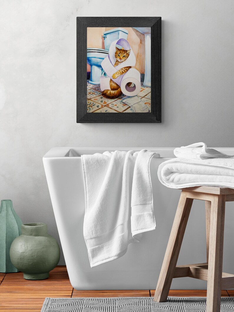 Peut inclure: Une peinture &agrave; l'aquarelle encadr&eacute;e repr&eacute;sente un chat roux envelopp&eacute; dans du papier toilette dans une salle de bain. La peinture est accroch&eacute;e au-dessus d'une baignoire blanche avec des serviettes blanches. Un tabouret en bois contient d'autres serviettes blanches.