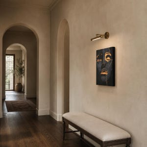 May include: A rectangular wall-mounted picture light in a brushed brass finish, featuring a slim cylindrical lamp head with an integrated LED strip, positioned above a vertical canvas painting depicting a dark face with gold leaf accents.