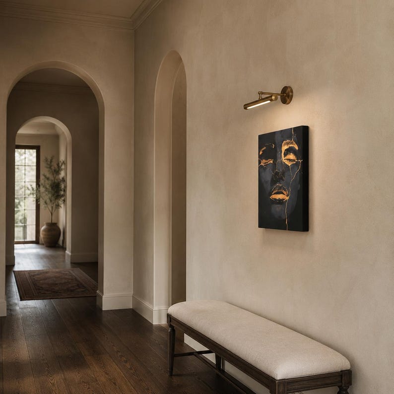 May include: A rectangular wall-mounted picture light in a brushed brass finish, featuring a slim cylindrical lamp head with an integrated LED strip, positioned above a vertical canvas painting depicting a dark face with gold leaf accents.