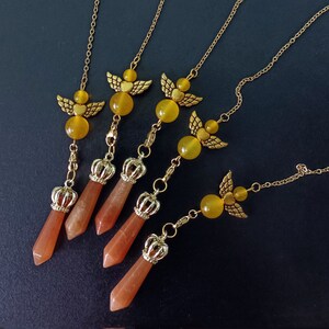 May include: Five gold-colored necklaces with angel wing pendants, yellow beads, and crown-topped orange crystal pendants. The necklaces are arranged on a dark surface, showcasing the jewelry's intricate details and design.