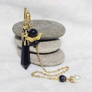 May include: A gold-toned pendulum with a dark blue, faceted crystal point and a decorative, ornate top. The pendulum is hanging from a gold chain with a pearl bead at the end.