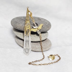 May include: A gold and clear crystal pendulum with a winged charm and a beaded chain. The pendulum is resting on a stack of gray stones.