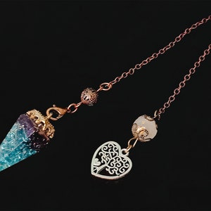 Puede incluir: Un péndulo de cristal azul y morado con un borde dorado y una cadena de cobre. La cadena tiene dos cuentas decorativas, una blanca y una dorada. El péndulo está unido a un dije de corazón plateado con un diseño de árbol.