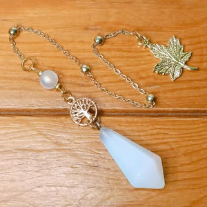 May include: A white crystal pendulum with a gold chain and a tree of life charm. The chain also has a gold maple leaf charm.