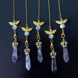 Puede incluir: Cinco cadenas doradas con dijes de alas de ángel y cuentas de cristal iridiscente, cada una terminando con una corona dorada y una punta de cristal morado.
