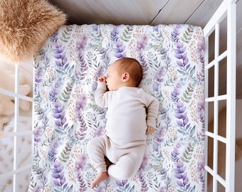 Op de afbeelding: Een baby slaapt vredig op een beddengoed met een aquarel bloemenpatroon. Het laken heeft paarse, lavendelkleurige en groene bladeren en bloemen op een witte achtergrond. De wieg is wit en er ligt een zacht kussen in de hoek.